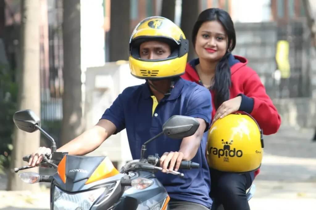 A man wearing a yellow helmet is riding a Rapido bike and a girl is sitting behind him holding a helmet in her hand.
